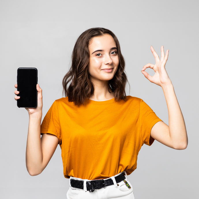 Smiling woman in orange shirt holding a smartphone and making an OK gesture on a gray background