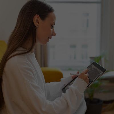 Woman sitting on a yellow couch using a tablet in a room with white walls and curtains.