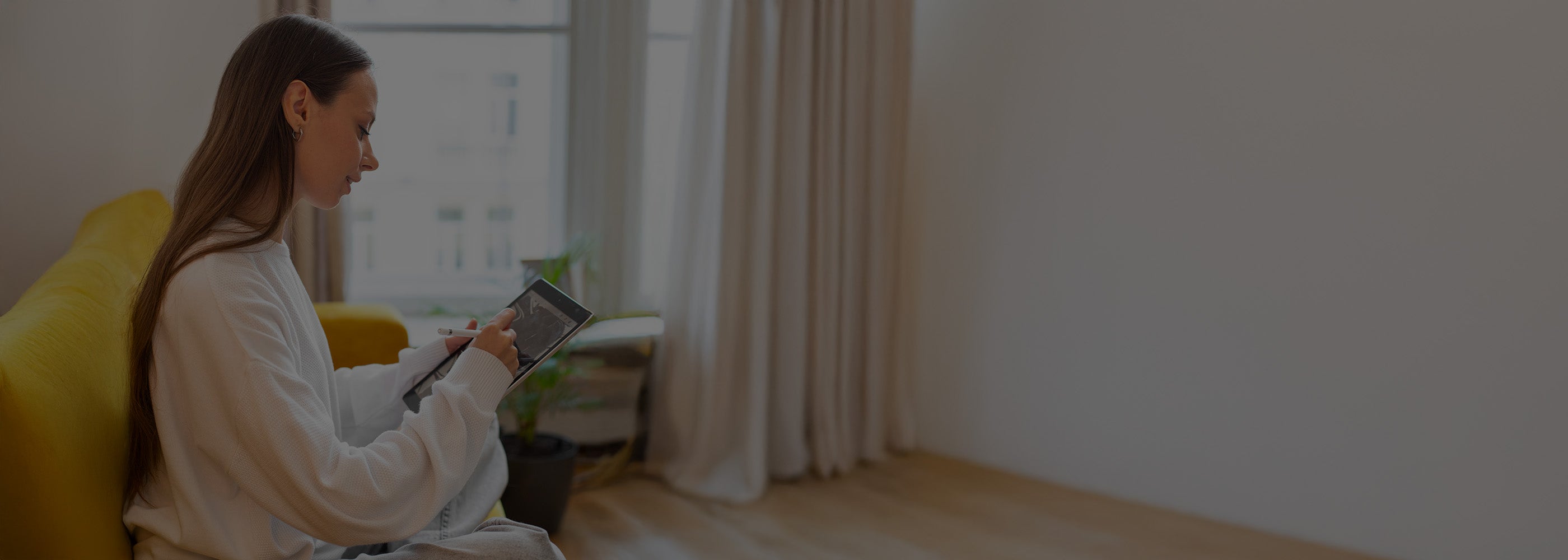 Woman sitting on a yellow couch using a tablet in a room with white walls and curtains.
