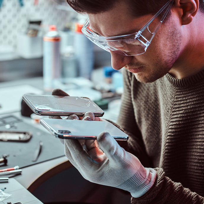 Person wearing safety glasses and gloves inspecting a smartphone in a workshop setting.
