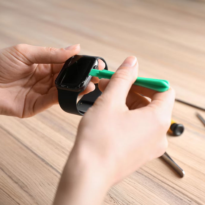Technician using a green plastic tool to repair a smartwatch on a wooden desk